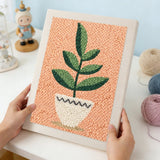 Hands holding a finished punch needle kit artwork featuring a green potted plant on a textured background.